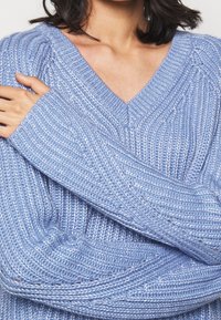 Světlomodrý pletený svetr s výstřihem do V, texturovaným žebrovaným vzorem a dlouhými širokými rukávy, vyrobený z měkké směsi vláken.