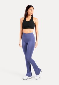 Reggiseno sportivo nero con scollo rotondo; leggings viola a vita alta in tessuto liscio; modello con indosso sneakers bianche.