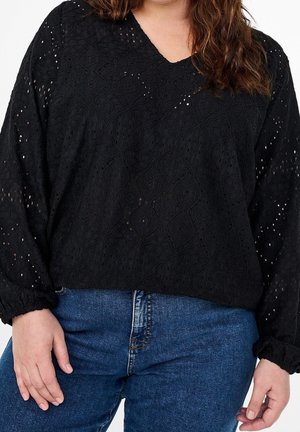 Persoon draagt een zwarte kanten top met lange mouwen en een V-hals, gecombineerd met blauwe spijkerbroek, staat met ontspannen handen bij de heupen.