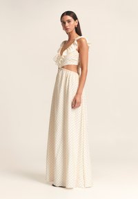 Zweiteiliger cremefarbener Anzug mit Polka-Dots. Das Oberteil ist mit Rüschen versehen und hat ein kurzes Design; der lange Rock verfügt über einen elastischen Bund für Komfort.