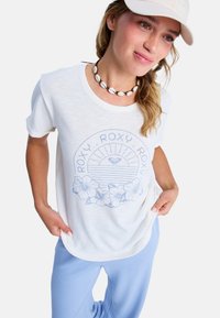 Witte t-shirt met een lichtblauwe grafische print van een zonnestraal en hibiscus bloemen, gecombineerd met lichtblauwe broek en een schelpen ketting.