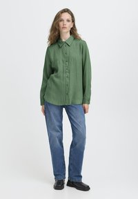 Femme aux cheveux ondulés portant une chemise à manches longues verte, un jean bleu et des chaussures noires, se tenant devant un fond uni.
