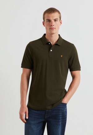 Giovane uomo che indossa una polo verde scuro a maniche corte e jeans blu, in piedi davanti a uno sfondo chiaro e semplice.