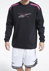 Persona vestida con sudadera negra con rayas rosas en los hombros y pantalones cortos de baloncesto blancos con diseño negro, de pie frente a un fondo blanco.