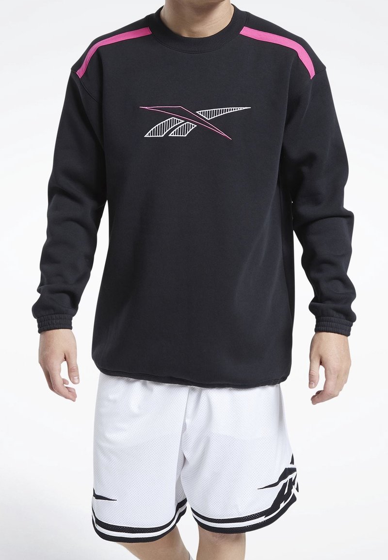 Persona vestida con sudadera negra con rayas rosas en los hombros y pantalones cortos de baloncesto blancos con diseño negro, de pie frente a un fondo blanco.