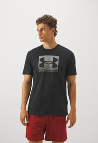 Svart t-skjorte med korte ermer som har en stor grå Under Armour-logo foran, laget av myk stoff med en klassisk passform.