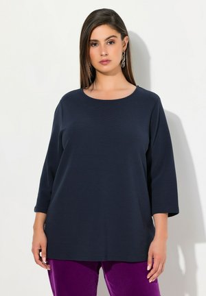 Haut à manches longues bleu marine avec un col rond et une coupe ample, confectionné dans un tissu texturé. Associé à un pantalon en velours violet.
