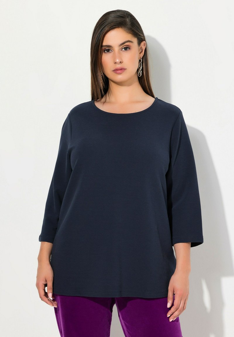 Marineblaue Langarmbluse mit Rundhalsausschnitt und lockerer Passform, aus strukturiertem Stoff. Kombiniert mit lila Samthose.