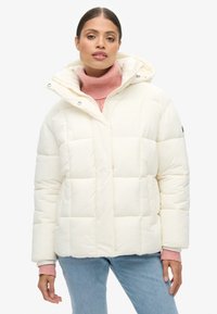 Chaqueta de plumas blanca con un diseño cuadrado, textura acolchada y capucha, combinada con un suéter de cuello alto rosa. Detalles de botones en el escote.