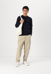 Μπλέ μακρυμάνικη μπλούζα με λαιμόκοψη και λογότυπο, μπεζ cargo παντελόνι με πλαϊνές τσέπες και λευκά αθλητικά παπούτσια. Το μοντέλο κρατάει μια καμβά τσάντα.