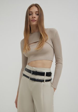 Top corto beige a coste con maniche lunghe e un dettaglio a taglio, abbinato a pantaloni beige a vita alta e cinture nere sovrapposte.