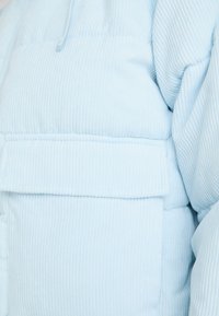 Svetle modrá kordurová péřová bunda s široce ribbed texturou, přední náprsní kapsou a vysokým límcem se zapínáním na knoflík.