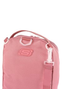 Bolso de tela rosa con superficie texturizada, herrajes transparentes, correa para el hombro y un logotipo prominente en la parte frontal. Diseño compacto.