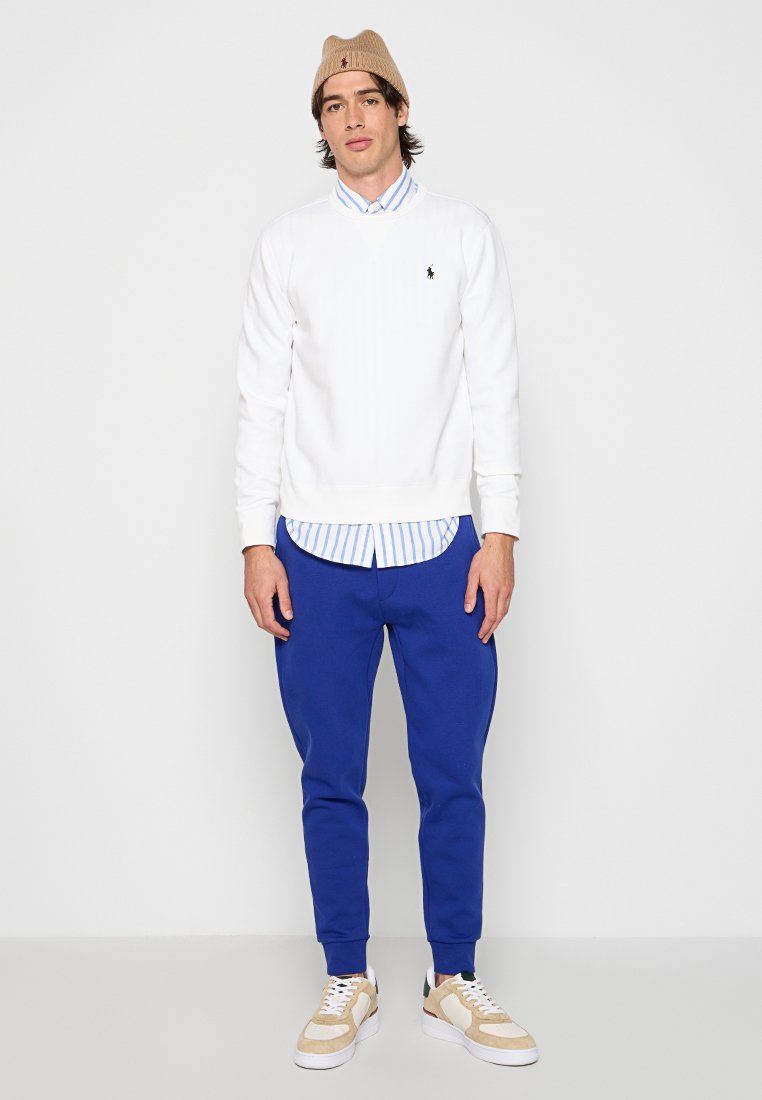 Biały sweter z prążkowanymi mankietami, założony na jasnoniebieską, w paski koszulę. Niebieskie joggery ze zwężanymi nogawkami i beżowe sneakersy dopełniają wygląd.