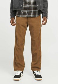 Pantalones de pana marrones con piernas anchas, que presentan una textura de estrías verticales. Combinados con una camisa de franela a cuadros en blanco y negro y zapatillas negras.