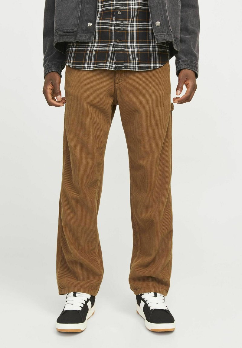 Pantalones de pana marrones con piernas anchas, que presentan una textura de estrías verticales. Combinados con una camisa de franela a cuadros en blanco y negro y zapatillas negras.