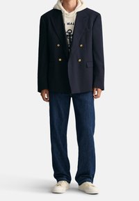 Granatowa marynarka dwurzędowa ze złotymi guzikami, połączona z jasną bluzą z kapturem, ciemnymi jeansami oraz kremowymi trampkami. Prosty design i dopasowanie.