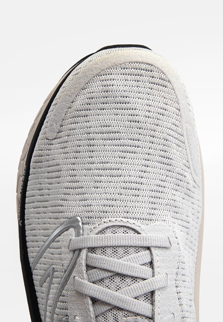 Zapatilla deportiva de color gris claro con una parte superior de tejido texturizado, cordones planos y una suela de goma negra. Presenta un sutil acento de logo en el costado.