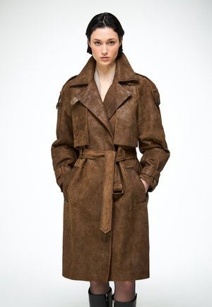 Donna con capelli corti e scuri che indossa un trench in camoscio marrone con cintura e stivali neri, in piedi davanti a uno sfondo bianco uniforme.