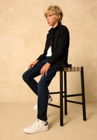 Cămașă neagră cu mânecă lungă peste un tricou alb, jeans din denim albastri și adidași albi. Stând pe un scaun de lemn pe un fundal bej.