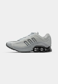 Gray athletic shoe featuring mesh and suede materials, curved shape, three black stripes, and a distinctive black shock-absorbing sole.