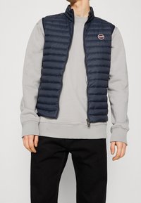 Navy puffer vest with horizontal stitching, high collar, and zippered pockets over a light gray sweatshirt. Features a brand logo on the chest.