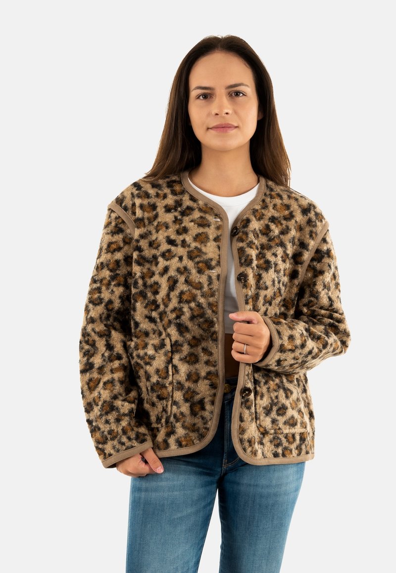Donna che indossa una giacca buttoned con stampa leopardo marrone sopra una maglietta bianca cropped e jeans blu, in piedi su uno sfondo semplice.
