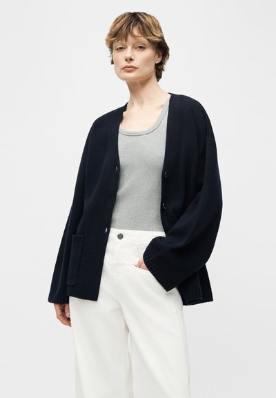 Femme portant un cardigan noir ample sur un débardeur gris et un pantalon large blanc, debout avec une main dans la poche devant un fond uni.