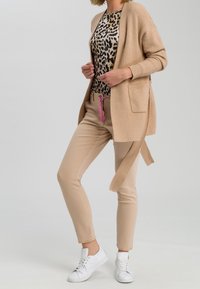 Kvinde iført beige strikcardigan, leopardmønstret top, beige bukser med lyserød snor, og hvide sneakers, stående mod grå baggrund.