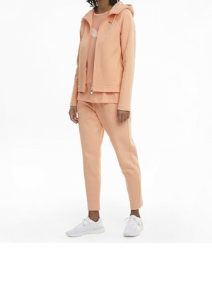 Perzikkleurige hoodie met rits en trekkoord, bijpassend perzikkleurig topje en taps toelopende joggingsbroek. Witte sneakers maken de outfit compleet.