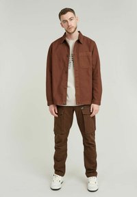 Bruine katoenen shirtjack met een borstzak, gecombineerd met bijpassende bruine cargo-broeken met rits- en klepzakken. Witte sneakers maken de outfit compleet.