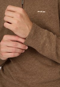 Pull en maille marron avec col zippé, poignets côtelés et un subtil motif blanc sur la poitrine. Texture douce et coupe décontractée.