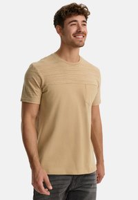 Beige kortærmet T-shirt med en tekstureret øverste del, rund halsudskæring og en lille brystlomme, lavet af blødt stof.