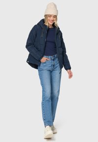 Mørkeblå dunjakke med hætte, sorte detaljer og elastiske manchetter; parret med lyseblå jeans og beige sneakers.