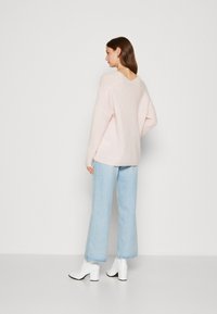 Lichtroze oversized trui met een V-back ontwerp, gecombineerd met lichtblauwe wijdvallende jeans en witte enkellaarsjes, weergegeven tegen een effen achtergrond.