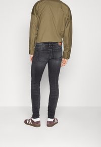 Jeans skinny gris oscuro con un lavado desgastado. Llevados con una camisa verde oliva y zapatos burdeos con rayas blancas.