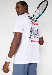 Hvid bomulds-t-shirt med et grafisk print på ryggen med "WANTED" i rødt, vestlig stil illustration og sorte accenter.