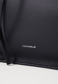 Borsa a tracolla nera in pelle texturizzata con un design elegante, caratterizzata da un sottile logo bianco "COCCINELLE" sul fronte e un aspetto minimalista.