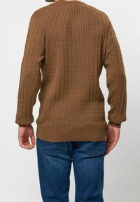 Pull en maille torsadée marron à manches longues, avec poignets et ourlet côtelés, porté sur un jean denim bleu. Présente un motif texturé et une coupe décontractée.