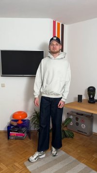 Pilkas didelio dydžio džemperis su kapišonu, tamsiai mėlynos plačiapėdės džinsinės kelnės ir sidabriniai sportiniai bateliai. Fone – augalai ir spalvingi sienų akcentai.