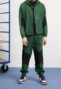 Giacca in nylon verde, leggera, con tasche laterali e rifiniture bianche. Abbinata a pantaloni da tuta verde e neri coordinati. Sneakers nere con accenti bianchi.