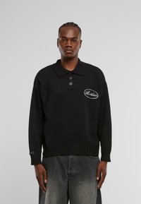 Schwarzer Strickpullover mit Polo-Kragen, ausgestattet mit drei Knöpfen und einem gestickten Logo auf der linken Brust. Ribbadapfen an den Ärmeln und am Saum.