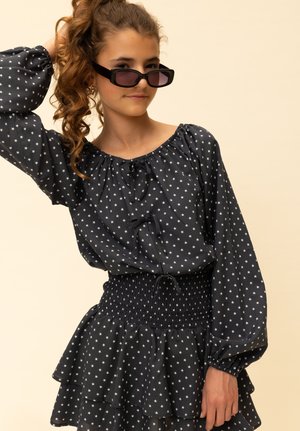 Zwart blouse met sterrenpatroon, pofmouwen, aantrekkoord bij de hals en een gerimpelde zoom, gecombineerd met een geplooide taille. Het model draagt rechthoekige zwarte zonnebrillen.