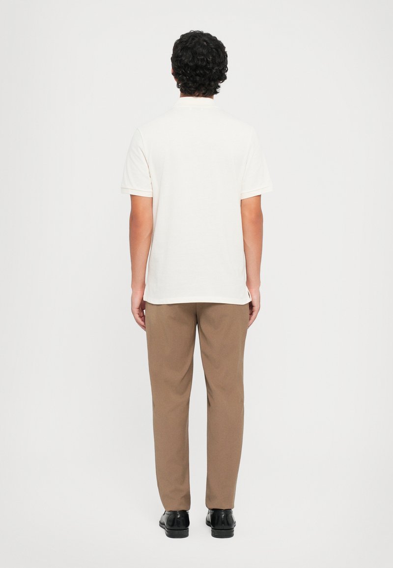 Helles, kurzärmliges Polohemd in Kombination mit braunen Hosen, von hinten betrachtet. Das Material wirkt glatt und hat ein einfaches Design.