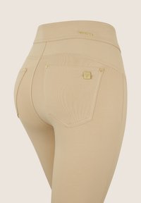 Leggings beige con vita alta, dotate di due tasche posteriori con dettagli in metallo dorato e un logo discreto sulla vita.