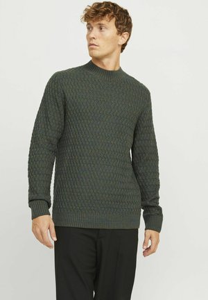 Suéter de punto en tonos verde oscuro y azul, con un patrón texturizado, cuello alto y puños y dobladillo de canalé. Combinado con pantalones negros.