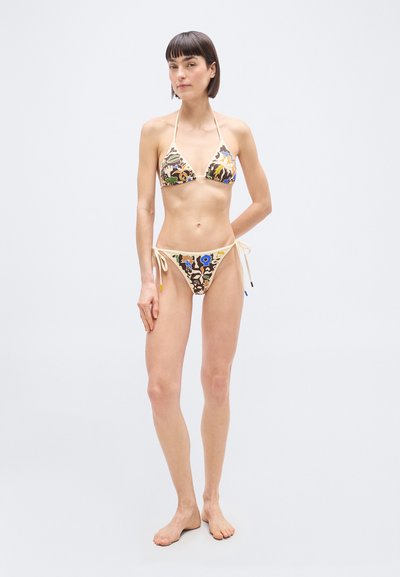 Γυναίκα που στέκεται ξυπόλητη φορώντας ένα μπικίνι δύο τεμαχίων με φλοράλ σχέδιο και κορδόνια, σε απλό ανοικτό φόντο.