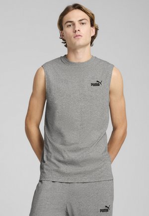Joven con camiseta sin mangas gris de Puma y pantalones cortos a juego, de pie con las manos detrás de la espalda frente a un fondo gris liso.