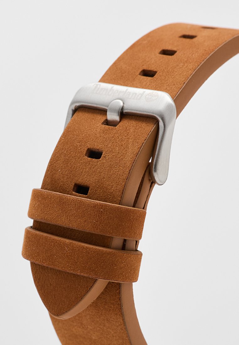 Correa de reloj de ante marrón con hebilla de metal plateada, que presenta varios agujeros de ajuste y elementos de diseño en capas para un ajuste seguro.