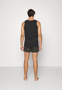 Camiseta sin mangas negra y pantalones cortos a rayas con una mezcla de verde oscuro y negro. Vista desde la parte de atrás, de pie descalzo contra un fondo claro.